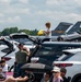 A crowded parking lot at what appears to be a military air show, with spectators gathered around their vehicles with open trunks to watch the event. A military jet aircraft tail fin is visible in the background, marked with 'AETC' and the serial number '66018', indicating it belongs to Air Education and Training Command of the U.S. Air Force. People are seated in folding chairs and standing between vehicles in warm, partly cloudy weather.