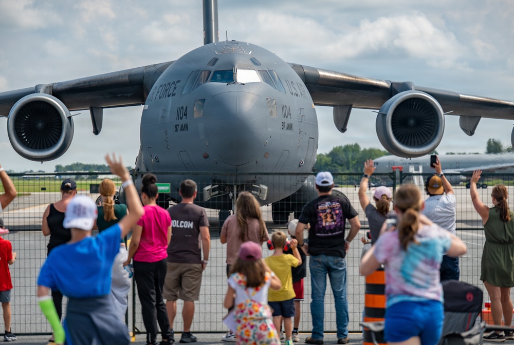 A crowd of spectators, including adults and children, wave at a U.S. Air Force C-17 Globemaster III cargo aircraft at what appears to be an air show. The aircraft bears the markings '1104' and '97 AMW' (Air Mobility Wing), with 'U.S. AIR FORCE' visible on the fuselage. The scene is set on an airfield tarmac with a security fence separating the crowd from the aircraft.