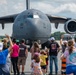 A crowd of spectators, including adults and children, wave at a U.S. Air Force C-17 Globemaster III cargo aircraft at what appears to be an air show. The aircraft bears the markings '1104' and '97 AMW' (Air Mobility Wing), with 'U.S. AIR FORCE' visible on the fuselage. The scene is set on an airfield tarmac with a security fence separating the crowd from the aircraft.
