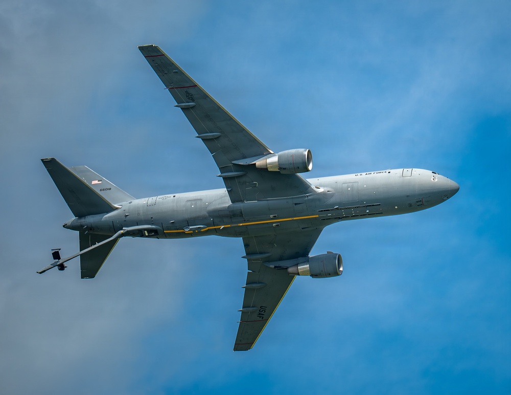 A U.S. Air Force military tanker aircraft is photographed from below against a partly cloudy blue sky. The aircraft is a KC-46 Pegasus (Boeing 767-based tanker) in standard gray military livery, with serial number 66018 visible on the fuselage. The image captures the underside of the aircraft in flight, showing its twin-engine configuration and aerial refueling boom extended beneath the tail.