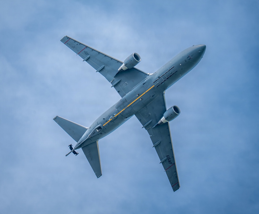 A United States Air Force (USAF) military aircraft is photographed from directly below as it flies overhead against a partly cloudy blue sky. The aircraft appears to be a KC-46 Pegasus or similar tanker/transport jet, identifiable by its gray military paint scheme, twin underwing engines, and a distinctive yellow stripe along the fuselage underside. The 'USAF' marking is clearly visible on the underside of the wing.