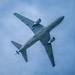 A United States Air Force (USAF) military aircraft is photographed from directly below as it flies overhead against a partly cloudy blue sky. The aircraft appears to be a KC-46 Pegasus or similar tanker/transport jet, identifiable by its gray military paint scheme, twin underwing engines, and a distinctive yellow stripe along the fuselage underside. The 'USAF' marking is clearly visible on the underside of the wing.