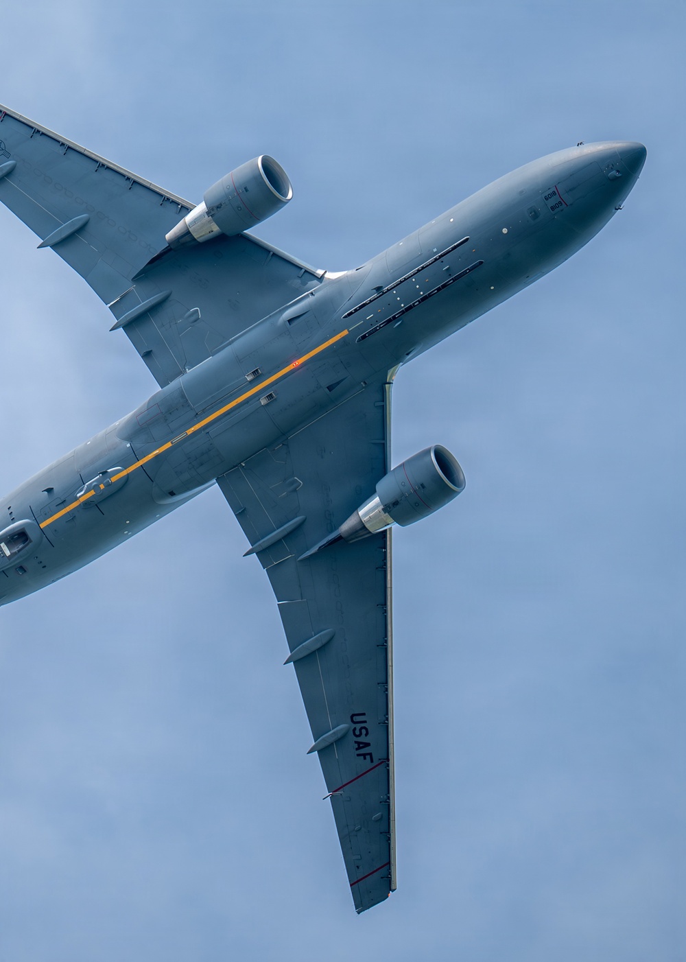 A United States Air Force (USAF) military aircraft, identifiable as a KC-46 Pegasus tanker, is photographed from directly below at a steep banking angle against a blue sky. The aircraft displays a gray paint scheme with a distinctive yellow/orange stripe along the fuselage and 'USAF' markings on the wing. The low-angle perspective provides a clear view of the underside of the wings, engines, and fuselage.