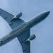 A United States Air Force (USAF) military aircraft, identifiable as a KC-46 Pegasus tanker, is photographed from directly below at a steep banking angle against a blue sky. The aircraft displays a gray paint scheme with a distinctive yellow/orange stripe along the fuselage and 'USAF' markings on the wing. The low-angle perspective provides a clear view of the underside of the wings, engines, and fuselage.