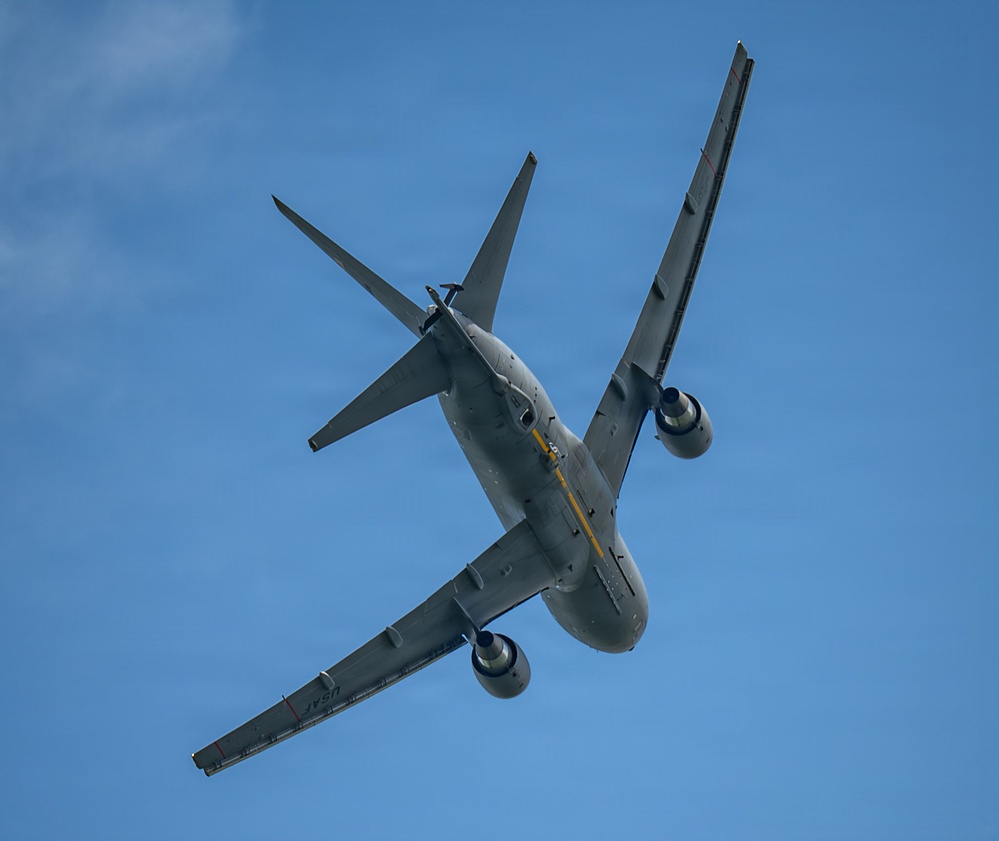 A military tanker aircraft, identifiable as a US Air Force KC-46 Pegasus or similar wide-body tanker, is photographed from below at a steep banking angle against a clear blue sky. The aircraft is painted in overall gray military livery and appears to be performing a maneuver, possibly during an airshow or training flight. The underside of the fuselage, wings, and engines are clearly visible, along with a yellow boom refueling system.