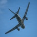 A military tanker aircraft, identifiable as a US Air Force KC-46 Pegasus or similar wide-body tanker, is photographed from below at a steep banking angle against a clear blue sky. The aircraft is painted in overall gray military livery and appears to be performing a maneuver, possibly during an airshow or training flight. The underside of the fuselage, wings, and engines are clearly visible, along with a yellow boom refueling system.