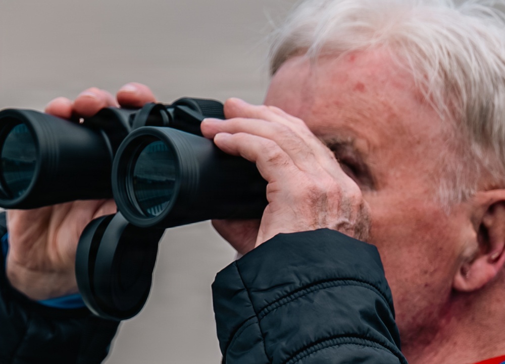 An elderly man with white hair is looking through a pair of black binoculars. He is wearing a dark navy quilted jacket and appears to be observing something in the distance. The image is a close-up side profile shot with a neutral, blurred background.