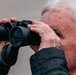 An elderly man with white hair is looking through a pair of black binoculars. He is wearing a dark navy quilted jacket and appears to be observing something in the distance. The image is a close-up side profile shot with a neutral, blurred background.