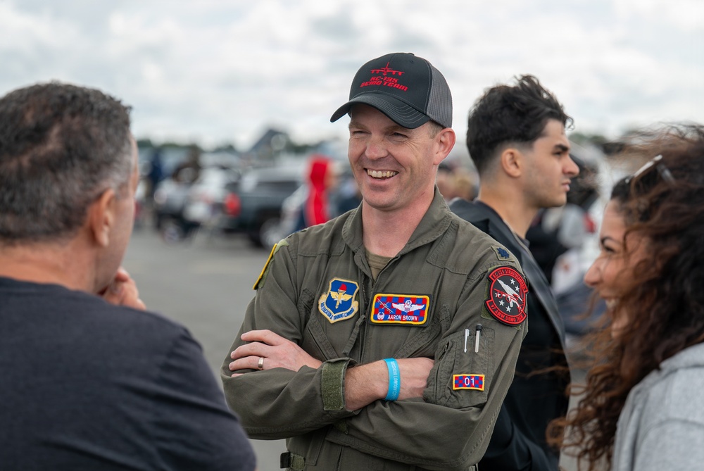 A smiling military aviator in an olive green flight suit, identified by a name tag as 'Aaron Brown,' stands with arms crossed at what appears to be an air show. He is wearing a black baseball cap with red text and multiple unit patches on his flight suit. He is engaged in conversation with several civilians in an outdoor setting with vehicles and crowds visible in the background.