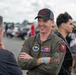 A smiling military aviator in an olive green flight suit, identified by a name tag as 'Aaron Brown,' stands with arms crossed at what appears to be an air show. He is wearing a black baseball cap with red text and multiple unit patches on his flight suit. He is engaged in conversation with several civilians in an outdoor setting with vehicles and crowds visible in the background.