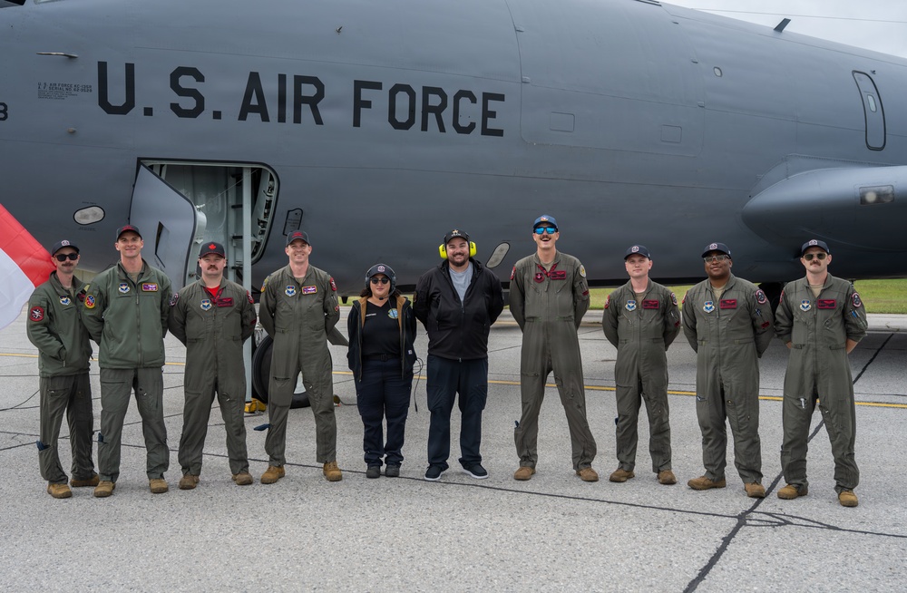 A group of nine individuals pose in front of a U.S. Air Force aircraft on a tarmac. Seven of the individuals are wearing olive green military flight suits, while two civilians stand among them. The group appears to be commemorating a visit or event associated with the aircraft.