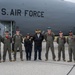 A group of nine individuals pose in front of a U.S. Air Force aircraft on a tarmac. Seven of the individuals are wearing olive green military flight suits, while two civilians stand among them. The group appears to be commemorating a visit or event associated with the aircraft.