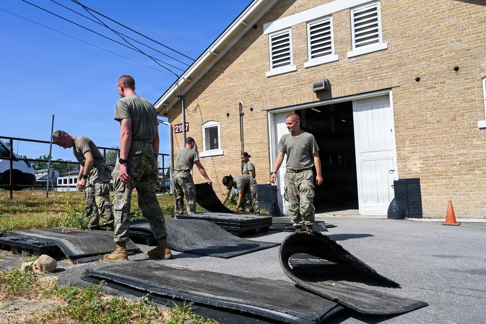 A group of military personnel in camouflage uniforms are working outdoors beside a tan brick building numbered '2187', handling large black rubber mats or panels. The soldiers appear to be moving, stacking, or organizing the heavy flat materials on a paved and grassy area. The scene suggests a maintenance, cleanup, or equipment organization task at a military facility.