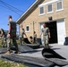 A group of military personnel in camouflage uniforms are working outdoors beside a tan brick building numbered '2187', handling large black rubber mats or panels. The soldiers appear to be moving, stacking, or organizing the heavy flat materials on a paved and grassy area. The scene suggests a maintenance, cleanup, or equipment organization task at a military facility.