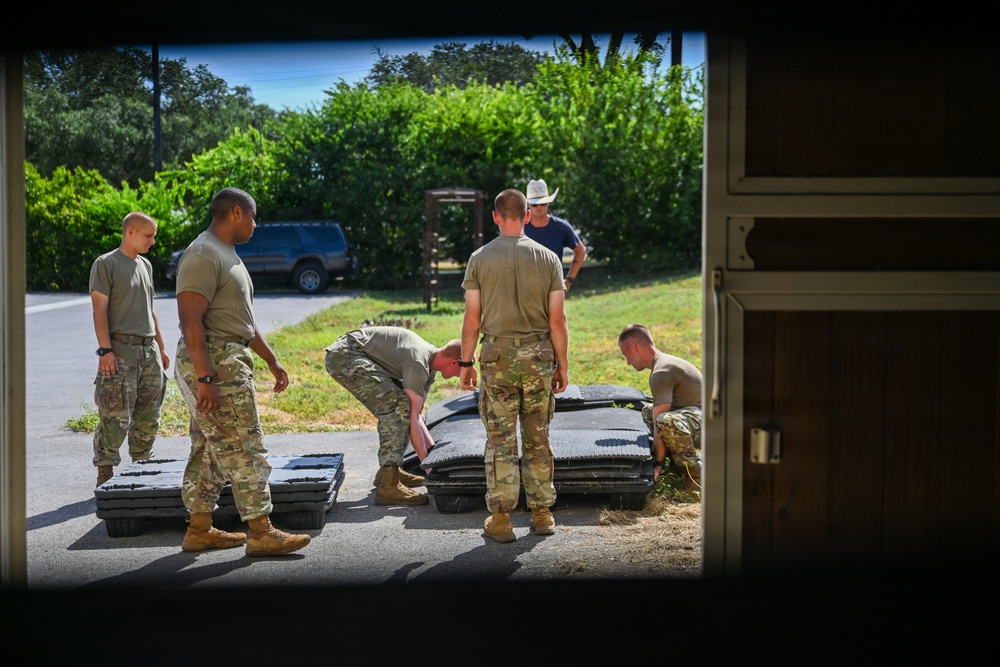 A group of military personnel in camouflage uniforms are gathered outdoors near what appears to be a building entrance or garage, working with large dark-colored equipment cases or panels on the ground. The scene is viewed through a doorway or window frame, suggesting the photographer is positioned inside a structure. A civilian in a cowboy hat and blue shirt observes the activity in the background.