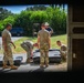 A group of military personnel in camouflage uniforms are gathered outdoors near what appears to be a building entrance or garage, working with large dark-colored equipment cases or panels on the ground. The scene is viewed through a doorway or window frame, suggesting the photographer is positioned inside a structure. A civilian in a cowboy hat and blue shirt observes the activity in the background.