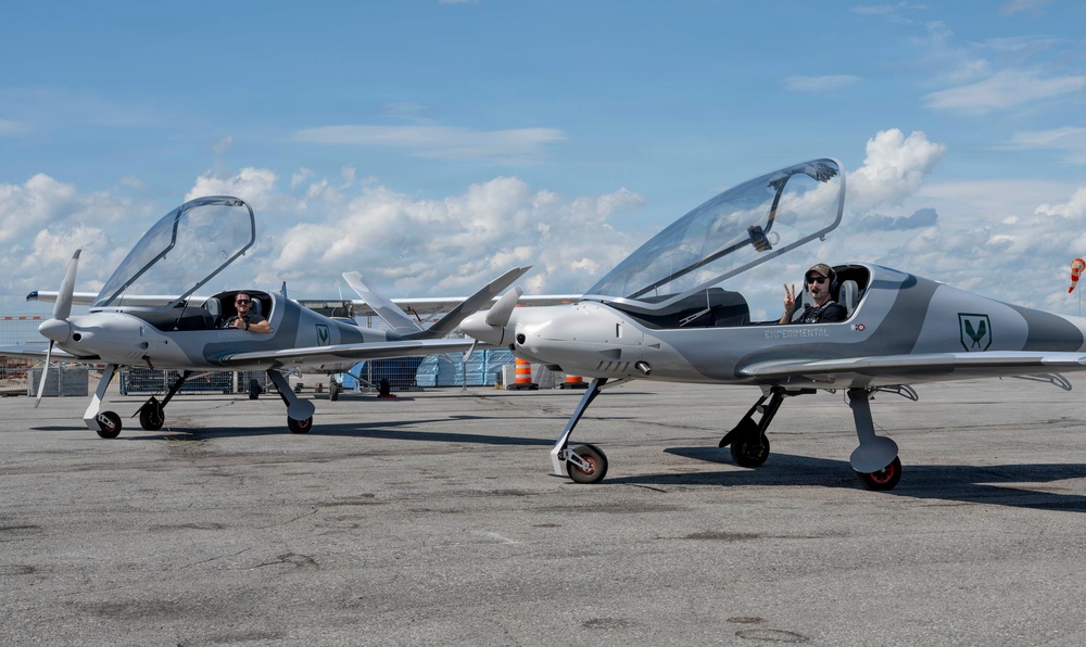 Two identical silver light aircraft are parked on an airport tarmac with their canopies open. Each aircraft has a pilot seated inside, one giving a thumbs-up and the other making a peace sign. The aircraft appear to be experimental or kit-built designs with a distinctive pusher or tractor propeller configuration and sleek aerodynamic fuselages.