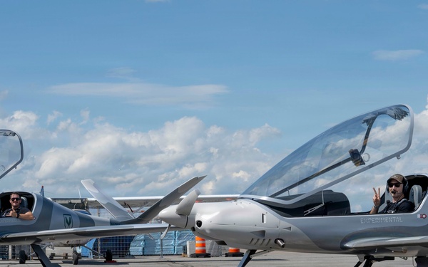Two identical silver light aircraft are parked on an airport tarmac with their canopies open. Each aircraft has a pilot seated inside, one giving a thumbs-up and the other making a peace sign. The aircraft appear to be experimental or kit-built designs with a distinctive pusher or tractor propeller configuration and sleek aerodynamic fuselages.