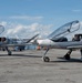 Two identical silver light aircraft are parked on an airport tarmac with their canopies open. Each aircraft has a pilot seated inside, one giving a thumbs-up and the other making a peace sign. The aircraft appear to be experimental or kit-built designs with a distinctive pusher or tractor propeller configuration and sleek aerodynamic fuselages.