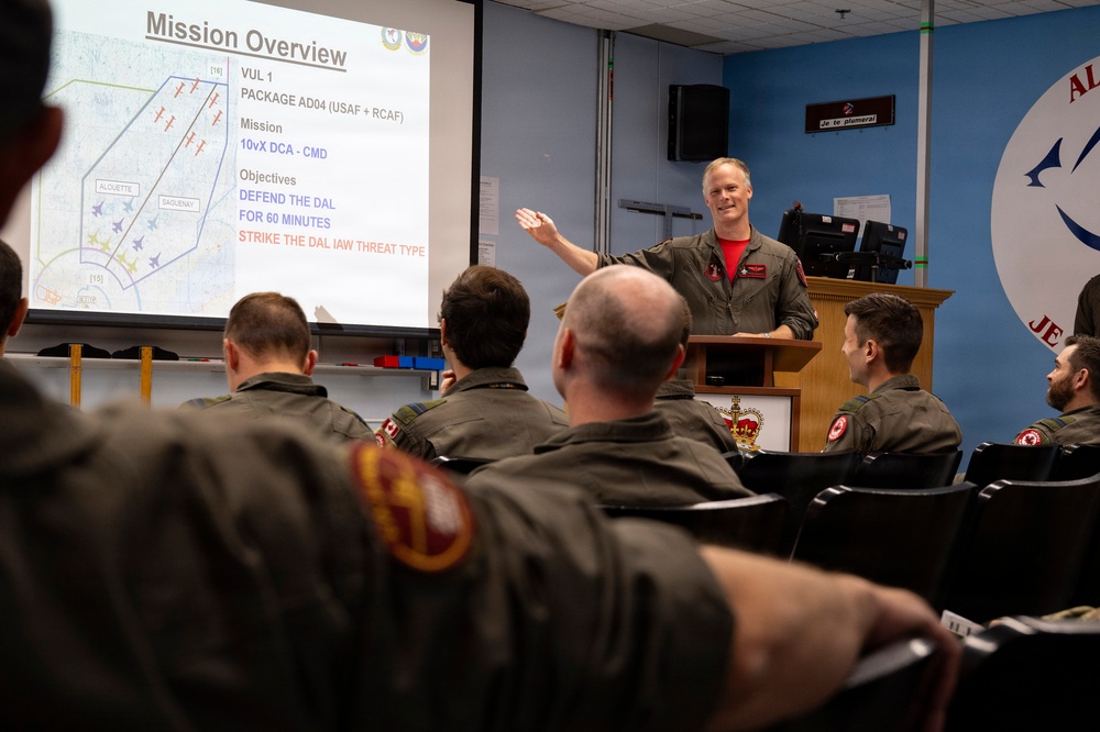 A military briefing room where a presenter in a flight suit is conducting a mission overview for a group of personnel. The slide displayed shows 'Mission Overview' with details about VUL 1, Package AD04 involving USAF and RCAF forces, with objectives including defending the DAL and striking targets. The audience consists of multiple personnel wearing olive/dark green flight suits with Canadian military patches.