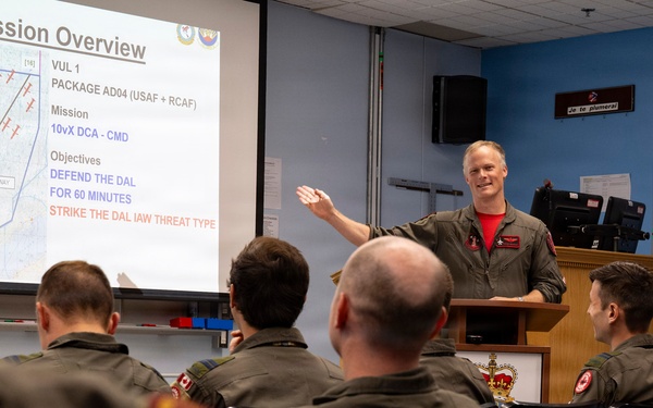 A military briefing room where a presenter in a flight suit is conducting a mission overview for a group of personnel. The slide displayed shows 'Mission Overview' with details about VUL 1, Package AD04 involving USAF and RCAF forces, with objectives including defending the DAL and striking targets. The audience consists of multiple personnel wearing olive/dark green flight suits with Canadian military patches.