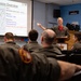 A military briefing room where a presenter in a flight suit is conducting a mission overview for a group of personnel. The slide displayed shows 'Mission Overview' with details about VUL 1, Package AD04 involving USAF and RCAF forces, with objectives including defending the DAL and striking targets. The audience consists of multiple personnel wearing olive/dark green flight suits with Canadian military patches.