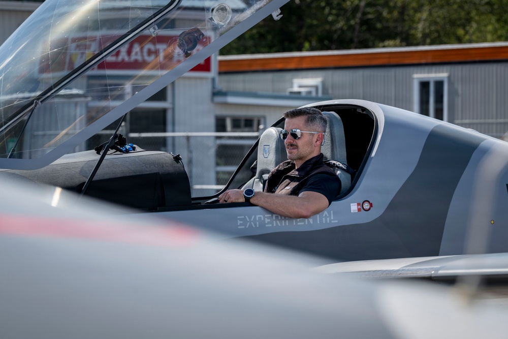 A man wearing aviator sunglasses and a tactical vest is seated in the open cockpit of a gray experimental aircraft, with the canopy raised. The aircraft bears the word 'EXPERIMENTAL' on its fuselage, indicating it is a homebuilt or prototype aircraft. The setting appears to be a small airfield or aviation facility, with buildings and signage visible in the background.