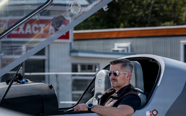 A man wearing aviator sunglasses and a tactical vest is seated in the open cockpit of a gray experimental aircraft, with the canopy raised. The aircraft bears the word 'EXPERIMENTAL' on its fuselage, indicating it is a homebuilt or prototype aircraft. The setting appears to be a small airfield or aviation facility, with buildings and signage visible in the background.