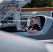 A man wearing aviator sunglasses and a tactical vest is seated in the open cockpit of a gray experimental aircraft, with the canopy raised. The aircraft bears the word 'EXPERIMENTAL' on its fuselage, indicating it is a homebuilt or prototype aircraft. The setting appears to be a small airfield or aviation facility, with buildings and signage visible in the background.