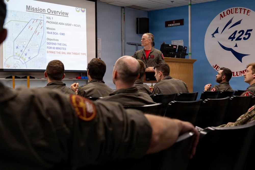 A military briefing is taking place in a room associated with the 425 'Alouette' Squadron, likely a Canadian Armed Forces unit. An instructor or senior officer is presenting a 'Mission Overview' slide to a group of personnel in flight suits. The briefing covers a joint USAF and RCAF package mission with defensive and strike objectives.