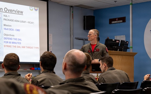 A military briefing is taking place in a room associated with the 425 'Alouette' Squadron, likely a Canadian Armed Forces unit. An instructor or senior officer is presenting a 'Mission Overview' slide to a group of personnel in flight suits. The briefing covers a joint USAF and RCAF package mission with defensive and strike objectives.