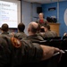 A military briefing is taking place in a room associated with the 425 'Alouette' Squadron, likely a Canadian Armed Forces unit. An instructor or senior officer is presenting a 'Mission Overview' slide to a group of personnel in flight suits. The briefing covers a joint USAF and RCAF package mission with defensive and strike objectives.