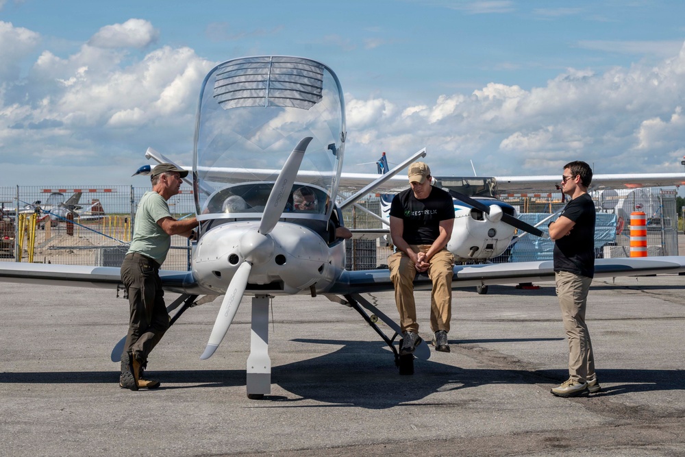 Three men are gathered around a small single-engine propeller aircraft on an airport tarmac. One man on the left is touching the nose of the aircraft, one is leaning against it in the center, and a third stands to the right. A fourth person appears to be seated inside the cockpit.