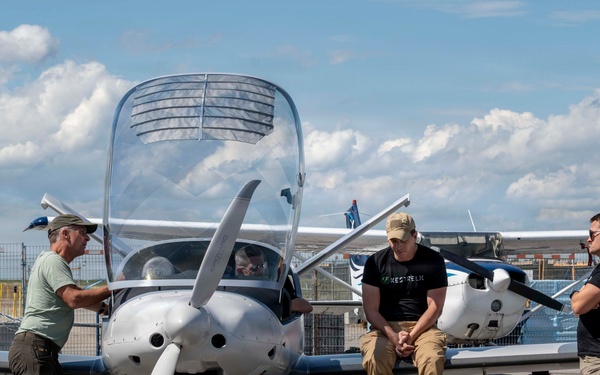 Three men are gathered around a small single-engine propeller aircraft on an airport tarmac. One man on the left is touching the nose of the aircraft, one is leaning against it in the center, and a third stands to the right. A fourth person appears to be seated inside the cockpit.
