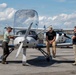 Three men are gathered around a small single-engine propeller aircraft on an airport tarmac. One man on the left is touching the nose of the aircraft, one is leaning against it in the center, and a third stands to the right. A fourth person appears to be seated inside the cockpit.