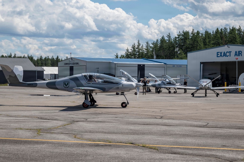 The image shows multiple sleek, modern light aircraft with gray camouflage-style paint schemes parked on an airport tarmac. Several people are visible near the aircraft, likely ground crew or pilots. The setting includes multiple aviation hangars in the background, with signage reading 'Exact Air' and 'Composite Aviation'.