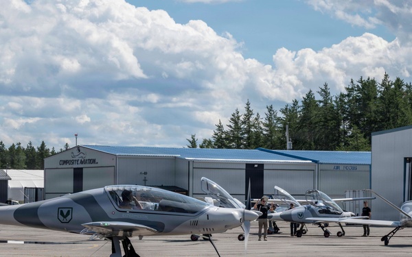 The image shows multiple sleek, modern light aircraft with gray camouflage-style paint schemes parked on an airport tarmac. Several people are visible near the aircraft, likely ground crew or pilots. The setting includes multiple aviation hangars in the background, with signage reading 'Exact Air' and 'Composite Aviation'.