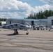 The image shows multiple sleek, modern light aircraft with gray camouflage-style paint schemes parked on an airport tarmac. Several people are visible near the aircraft, likely ground crew or pilots. The setting includes multiple aviation hangars in the background, with signage reading 'Exact Air' and 'Composite Aviation'.