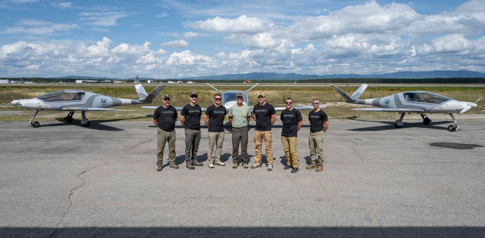 A group of seven men pose on an airport tarmac in front of two small aircraft, most wearing matching black t-shirts with a logo. The aircraft appear to be light sport or experimental planes with a distinctive modern design, featuring a green 'V' logo on the tail. The setting appears to be a regional airport with mountains and open sky visible in the background.