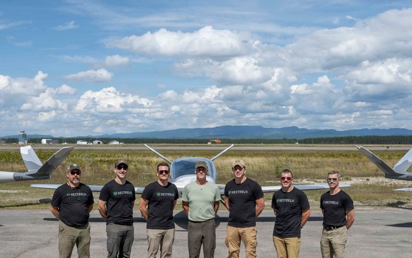 A group of seven men pose on an airport tarmac in front of two small aircraft, most wearing matching black t-shirts with a logo. The aircraft appear to be light sport or experimental planes with a distinctive modern design, featuring a green 'V' logo on the tail. The setting appears to be a regional airport with mountains and open sky visible in the background.