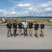 A group of seven men pose on an airport tarmac in front of two small aircraft, most wearing matching black t-shirts with a logo. The aircraft appear to be light sport or experimental planes with a distinctive modern design, featuring a green 'V' logo on the tail. The setting appears to be a regional airport with mountains and open sky visible in the background.