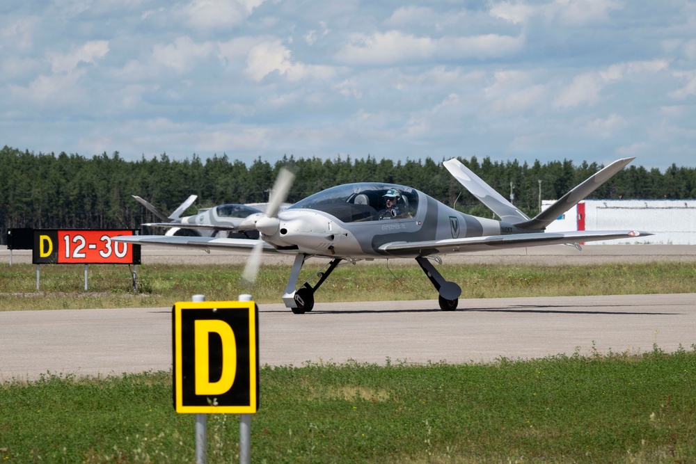 A sleek, modern light aircraft with a spinning propeller is taxiing on an airport taxiway near taxiway signage. The aircraft appears to be a canard-style or composite design in gray and white livery with a visible pilot in the cockpit. A second similar aircraft is visible in the background, suggesting a formation or fleet operation.