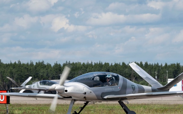 A sleek, modern light aircraft with a spinning propeller is taxiing on an airport taxiway near taxiway signage. The aircraft appears to be a canard-style or composite design in gray and white livery with a visible pilot in the cockpit. A second similar aircraft is visible in the background, suggesting a formation or fleet operation.