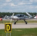 A sleek, modern light aircraft with a spinning propeller is taxiing on an airport taxiway near taxiway signage. The aircraft appears to be a canard-style or composite design in gray and white livery with a visible pilot in the cockpit. A second similar aircraft is visible in the background, suggesting a formation or fleet operation.