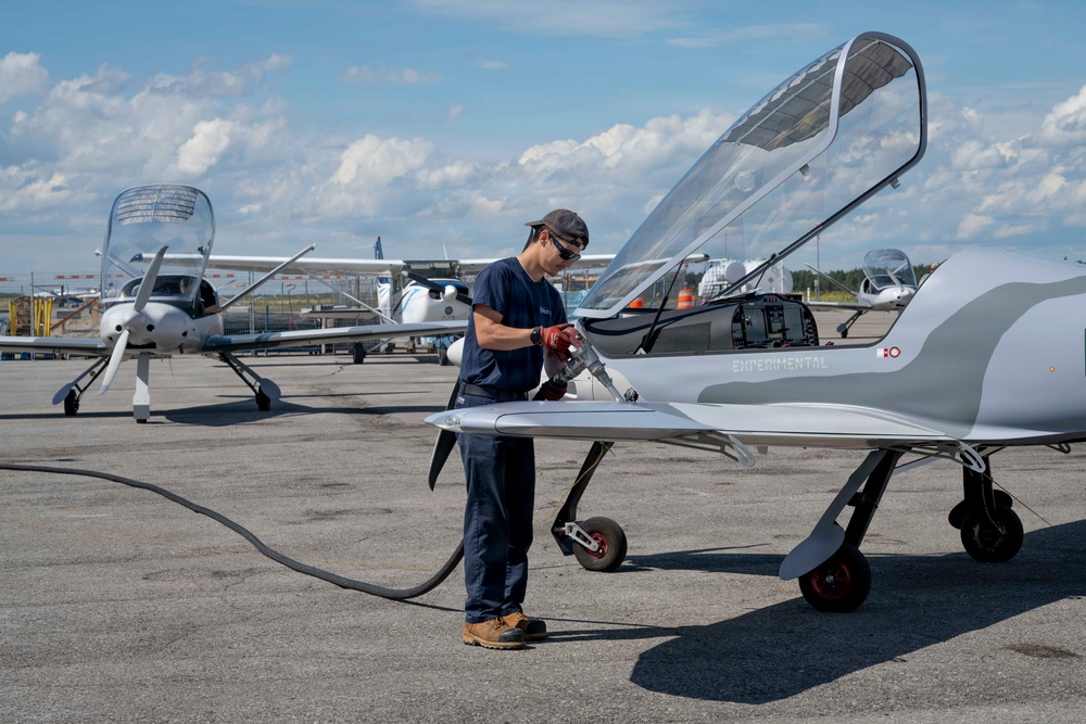 A technician in navy blue work clothes is fueling or servicing a sleek, low-profile experimental aircraft on an airport tarmac. The aircraft is labeled 'EXPERIMENTAL' on its fuselage and features a raised bubble canopy, suggesting it is a prototype or homebuilt design. Several other general aviation aircraft are visible in the background under a partly cloudy sky.