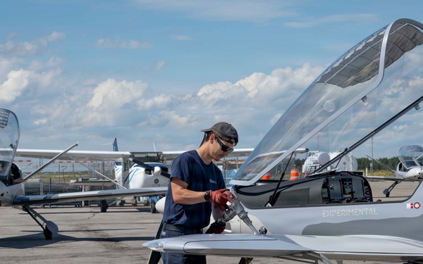 A technician in navy blue work clothes is fueling or servicing a sleek, low-profile experimental aircraft on an airport tarmac. The aircraft is labeled 'EXPERIMENTAL' on its fuselage and features a raised bubble canopy, suggesting it is a prototype or homebuilt design. Several other general aviation aircraft are visible in the background under a partly cloudy sky.