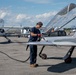 A technician in navy blue work clothes is fueling or servicing a sleek, low-profile experimental aircraft on an airport tarmac. The aircraft is labeled 'EXPERIMENTAL' on its fuselage and features a raised bubble canopy, suggesting it is a prototype or homebuilt design. Several other general aviation aircraft are visible in the background under a partly cloudy sky.