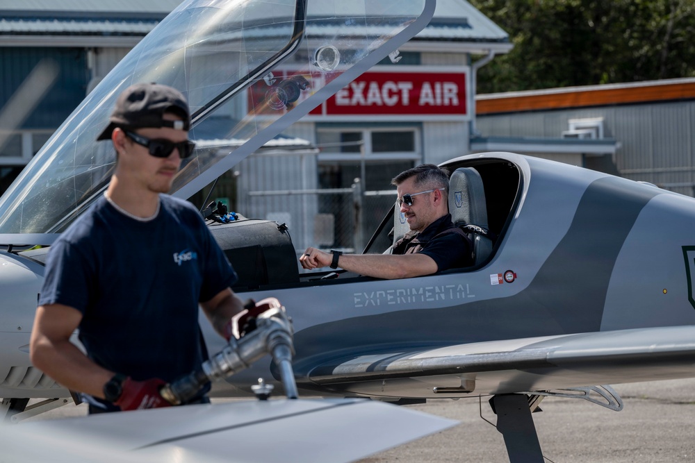 Two men are at a small airfield near an experimental aircraft, with one person seated in the cockpit and another standing outside holding a fuel nozzle. The aircraft is labeled 'EXPERIMENTAL' on its fuselage and appears to be a sleek, modern light aircraft. A building with a red sign reading 'EXACT AIR' is visible in the background.