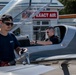 Two men are at a small airfield near an experimental aircraft, with one person seated in the cockpit and another standing outside holding a fuel nozzle. The aircraft is labeled 'EXPERIMENTAL' on its fuselage and appears to be a sleek, modern light aircraft. A building with a red sign reading 'EXACT AIR' is visible in the background.