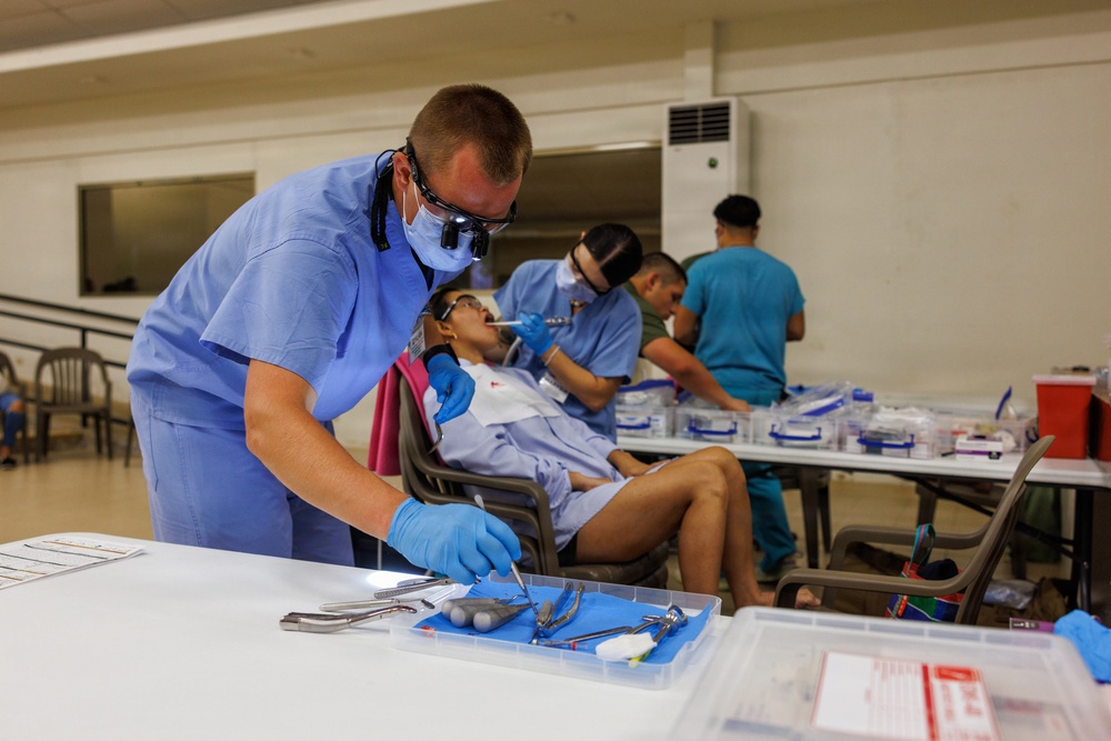 A field or community dental clinic is in operation inside a large indoor hall or gymnasium. Multiple dental practitioners in scrubs and protective gear are simultaneously treating patients seated in portable chairs. The setting appears to be a temporary or mobile dental outreach event with supplies organized on folding tables.